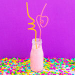 Party pink cocktail in bottle with drinking straws on purple background with confetti
