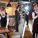 Manager and young waiters working together and preparing restaurant for opening: bearded man wiping wooden table, handsome brunette man cleaning wineglass