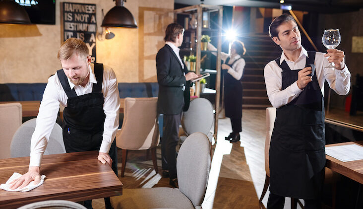 Manager and young waiters working together and preparing restaurant for opening: bearded man wiping wooden table, handsome brunette man cleaning wineglass