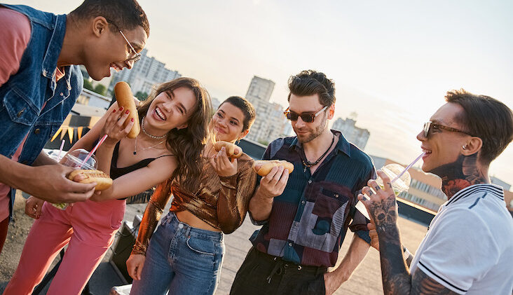 cheerful multicultural people in vivid outfits enjoying delicious hot dogs on rooftop at party